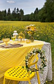 Blumenkranz auf gelbem Metallstuhl vor Tisch mit gelben Farbakzenten, im Hintergrund Rapsfeld