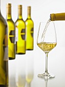 Weißwein wird in ein Weißweinglas eingeschenkt, eine Reihe Weinflaschen im Hintergrund