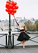 A young woman on a bridge wearing a black petticoat dress with a pink belt and holding a bunch of red balloons