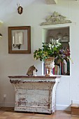 Vintage Konsolentisch mit abblätternder Farbe, darauf antikes Pflanzgefäss mit Blätterzweigen vor Nische in Wand, seitlich Wandkerzenhalter in leerem Bilderrahmen