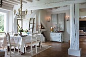 White chairs with carved backrests, table with white tablecloth and whitewashed supporting structure in dining area of old country house next to window