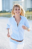 Junge blonde Frau in hellblauem Hemd und weisser Hose beim Joggen am Strand