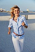 A young blonde woman jogging along a beach wearing a light blue shirt and white trousers