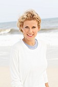 A blonde woman on a beach wearing a blue top and a white round-neck jumper