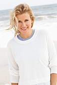 A blonde woman on a beach wearing a blue top and a white round-neck jumper