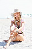 Junge blonde Frau mit Sommerhut, Schal und rosa T-Shirt sitzt im Sand am Strand