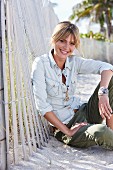 A young blonde woman sitting in the sand wearing a denim blouse and green trousers