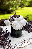 A jar of homemade elderberry jelly with a decorative lived and a sprig of ripe elderberries on a garden table