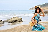 Frau in türkis gemustertem Kleid und Strohhut kniet am Strand