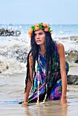 Frau in bunt gemustertertem Kleid und Blumenkranz kniet am Strand