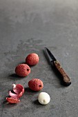 Lychees, whole and peeled, on a grey stone surface with a knife