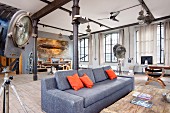 Graues Polstersofa mit orangefarbenen Kissen flankiert von Scheinwerfer-Stehleuchten in Loft-Wohnung mit schwarzen Metallstützen