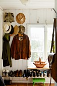 Garderobenecke mit aufgehängten Jacken und Hüten an weisser Holzwand, vollgestellte Schuhablage unter Fenster