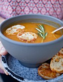A woman holding a bowl of roasted vegetable soup garnished with rosemary and cheese crostini