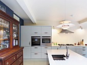 Antique, glass-fronted cabinet in open-plan, white kitchen with designer pendant lamp