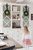 Girl walking past white kitchen dresser decorated with wreaths of box leaves