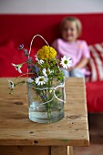 Wild flowers in glass of water on wooden table; little girl sitting on sofa in background
