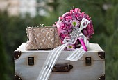 Romantischer pinkfarbener Hochzeitsstrauß mit weißem Schleifenband und Brosche neben elegantem Täschchen auf Vintagekoffer