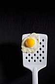 A fried quail's egg on a spatula on a black surface