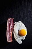 A fried egg and a rash of bacon on a dark surface (seen from above)