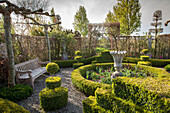 Garden bench, stone urn and beds of tulips inside circular hedge surrounded by gravel patch and clipped box bushes