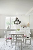 White table and wooden chairs below chandelier in rustic interior with white wooden floor