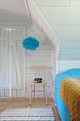 White, wood-clad attic bedroom with pink-painted chair below blue pompom suspended from ceiling