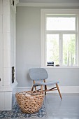Stuhl mit hellgrauem Polsterbezug vor Fenster in Zimmerecke, im Vordergrund Weidenkorb mit Brennholz vor Kamin
