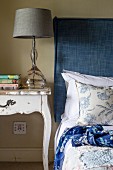 Table lamp with grey lampshade on vintage bedside table next to bed with headboard upholstered in blue fabric