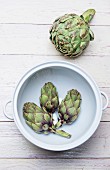 A fresh artichoke and baby artichokes in a porcelain bowl on the white wooden surface