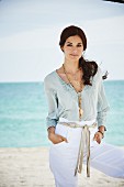 A brunette woman on a beach wearing white trousers and a light tunic with lace detailing