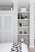 View over bed of shelves in narrow niche and fitted wardrobe with white louvre doors