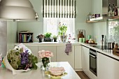 weiße Einbauküche mit Blumenstrauss auf Tisch, Übereck-Küchenzeile vor Fenster mit Küchenutensilien