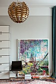 Modernes Bild an hellgraue Wand gelehnt, davor Vintage Drehstuhl und glänzende Pendelleuchte