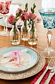 Romantic place setting with bird motif on plate and coloured crystal glasses next to roses in glass bottles