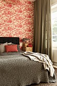 Blanket on double bed with headboard against wall with red and white toile de jouy wallpaper