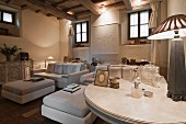 Im Vordergrund Tischleuchte auf rundem Tisch, elegante, weiße Sofagarnitur mit passenden Couchtischen in renoviertem, rustikalem Wohnzimmer