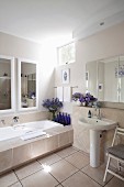 Badewanne mit breiter, gefliesten Ablage, Wandspiegel, blaue Flaschen und Blumensträusse