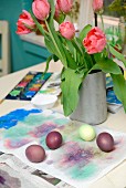 Painted Easter eggs on paper in front of metal vase of pink tulips