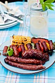A barbecue platter with a grilled corn cob, a sausage skewer and sausages
