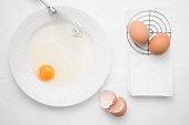 A broken egg on a plate with a whisk next whole eggs and egg shells