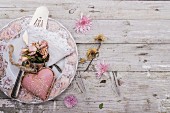 Rosa Dekoherz und getrocknetes Rosensträuschen auf nostalgischem Teller mit verstreuten Blumenblüten auf rustikaler Holzunterlage