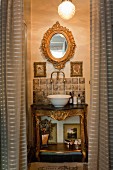 Baroque-style washstand with gilt details and white china basin below oval, gilt-framed mirror seen through open curtains in doorway