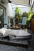 Floor cushions and vintage chairs with pale blue upholstery around fire bowl on balcony with pebble floor