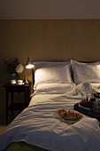 Brennende Nachttischlampe neben Doppelbett mit weisser Bettwäsche, im Vordergrund Frühstückstablett
