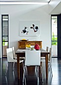 White modern chairs around wooden dining table in front of artwork above chest of drawers in background