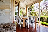 White wicker chairs at wooden table on reddish wooden floor of veranda with white wooden pillars