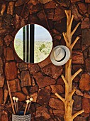 Round mirror next to hat hung from branch coat rack on rustic, rough stone wall