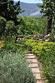 Schmaler Plattenweg im Garten, im Hintergrund Rosenbüsche zwischen geschnittenen Hecken und offene Berglandschaft