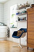 Retro swivel chair below crockery on String shelves next to wooden cabinet with roller door in kitchen area